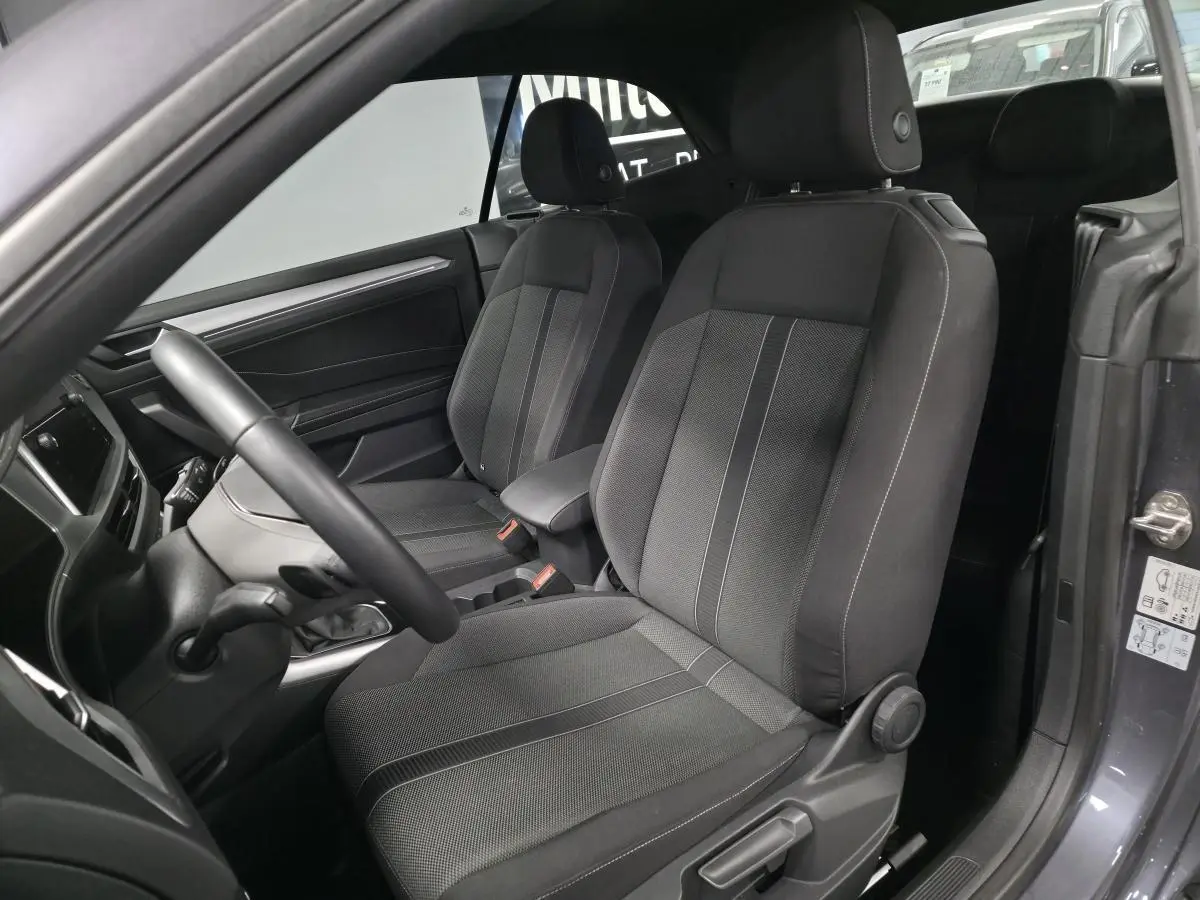 Vue intérieure côté conducteur du Volkswagen T-Roc 2024 gris, sièges tissu noir avec accoudoir central et volant multifonction.