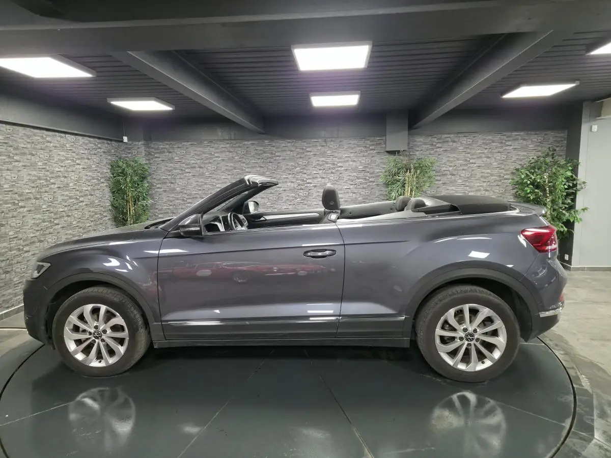 Profil droit d'un Volkswagen T-Roc cabriolet gris métallisé 2024 avec capote noire repliée en intérieur showroom.