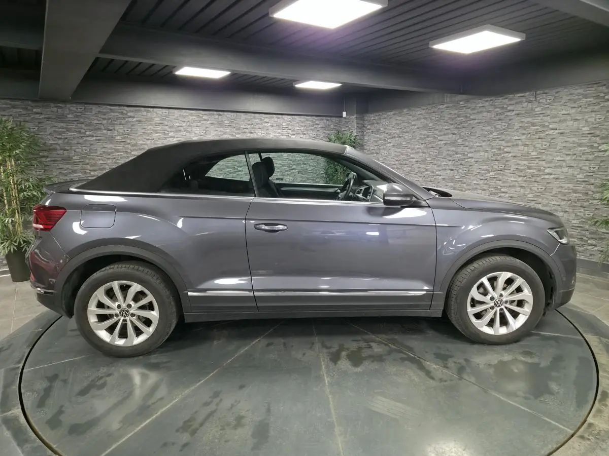 Volkswagen T-Roc gris métallisé vue de profil côté gauche, capote noire textile fermée en intérieur showroom.