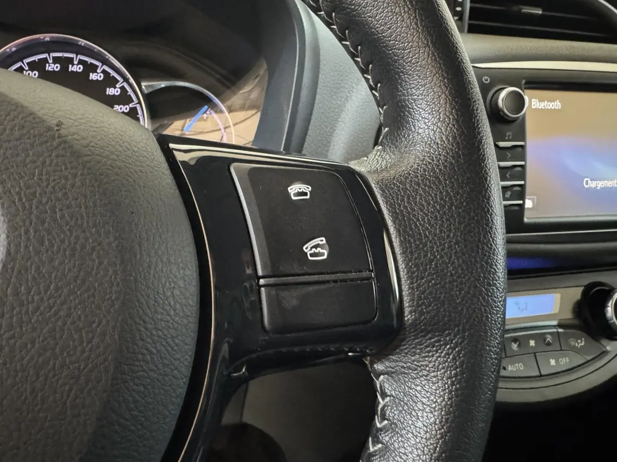 Gros plan sur le volant cuir noir de la Toyota Yaris hybride 2019, avec commandes téléphoniques et écran tactile Bluetooth visible.