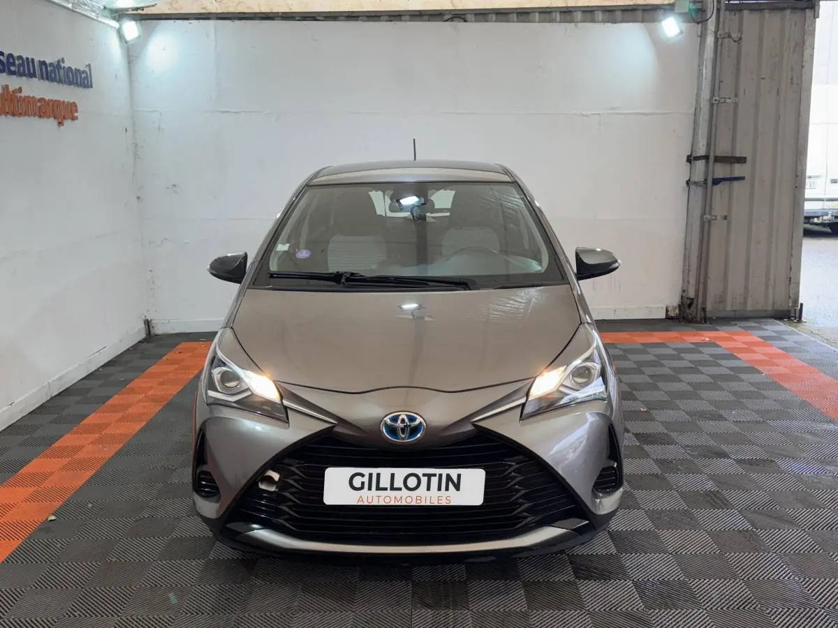 Vue avant d'une Toyota Yaris hybride gris foncé avec phares allumés dans un garage éclairé.