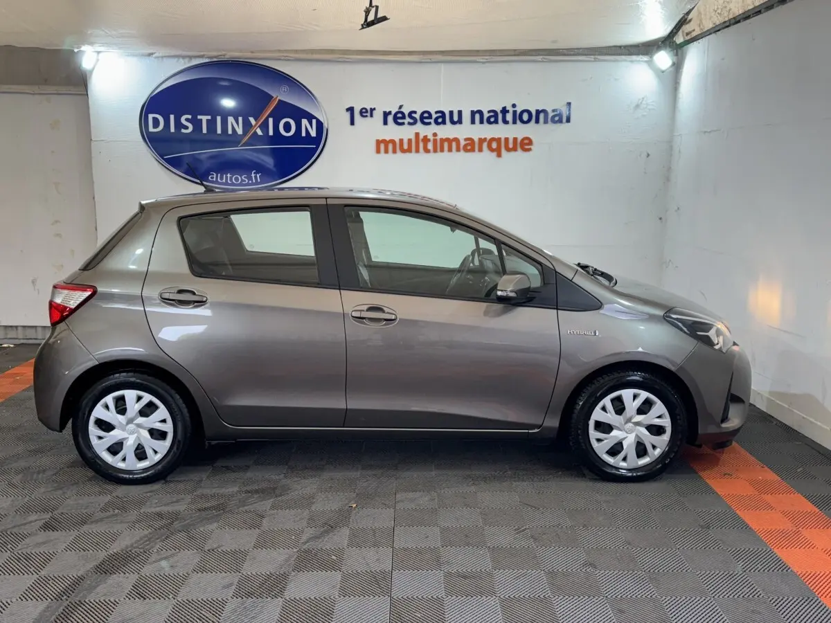 Vue latérale droite d'une Toyota Yaris hybride gris foncé, avec jantes blanches et badge Hybrid visible sur l'aile avant.