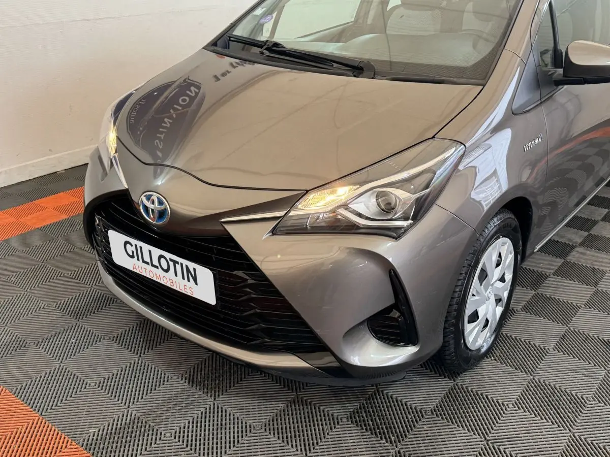Vue 3/4 avant droit d'une Toyota Yaris hybride gris foncé avec phares allumés et jantes acier.