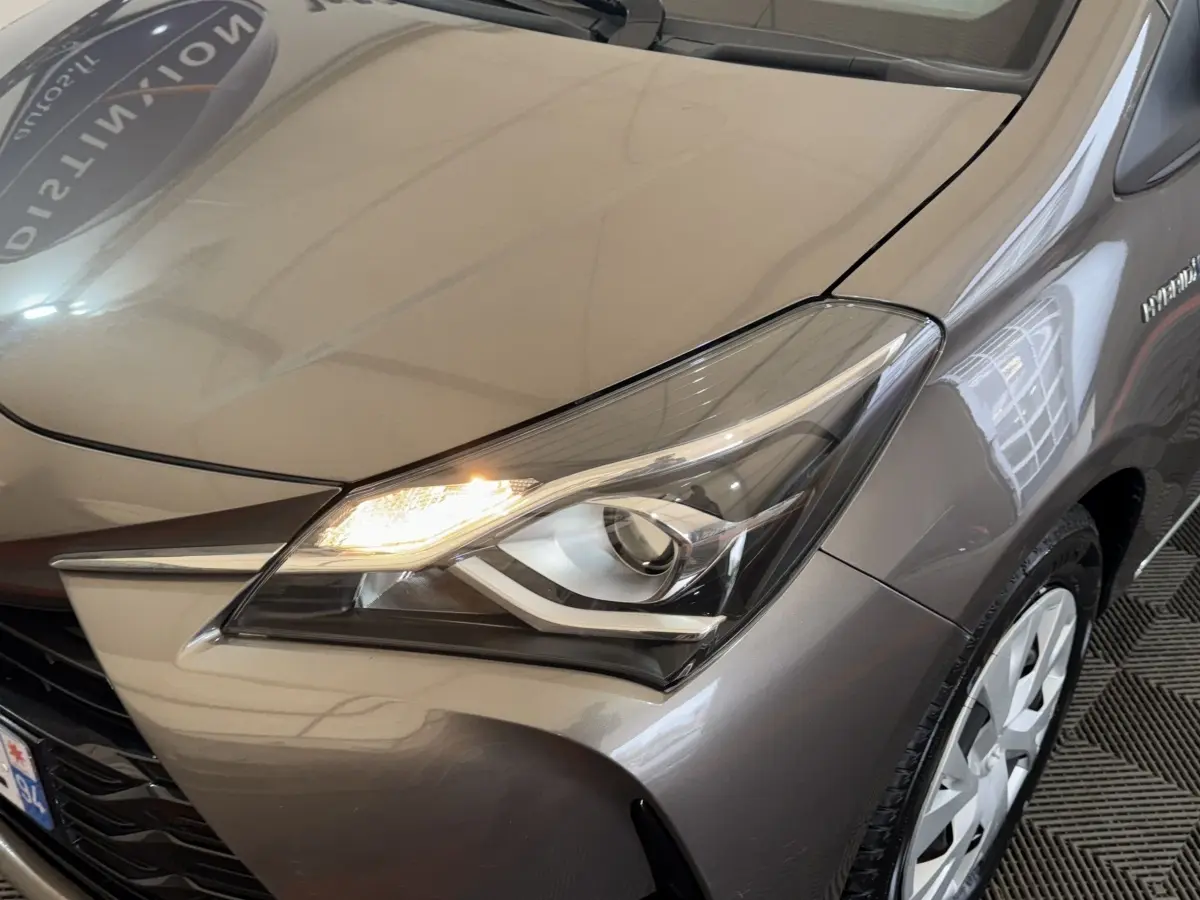 Gros plan sur l'avant droit gris foncé d'une Toyota Yaris 1.5 VVT-I Hybrid 2019 avec phare allumé et jante visible.