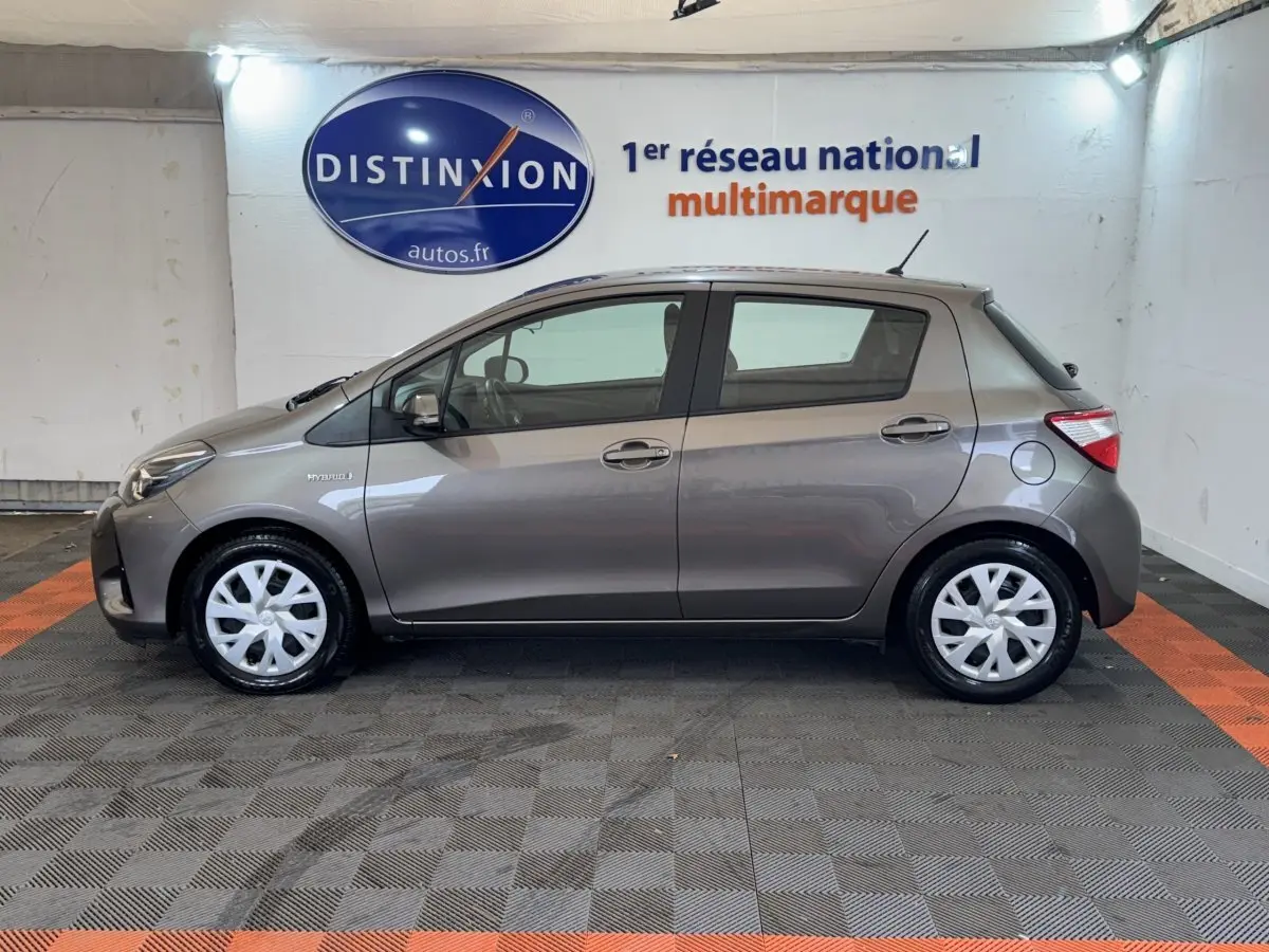 Vue de profil côté gauche d'une Toyota Yaris hybride gris foncé avec jantes blanches en intérieur showroom.