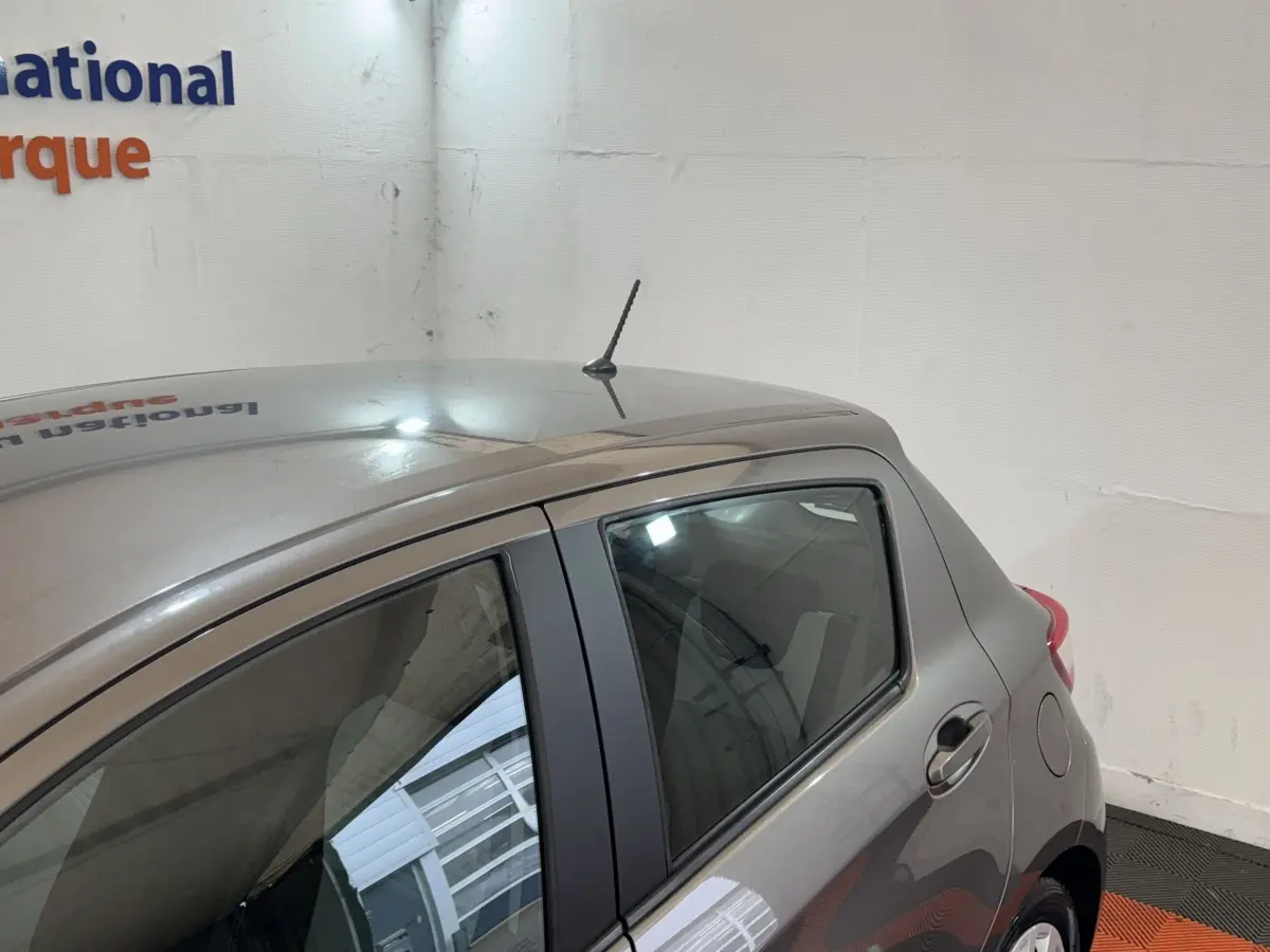 Vue latérale arrière droite d'une Toyota Yaris 1.5 VVT-I Hybrid gris foncé en intérieur, avec antenne de toit visible.