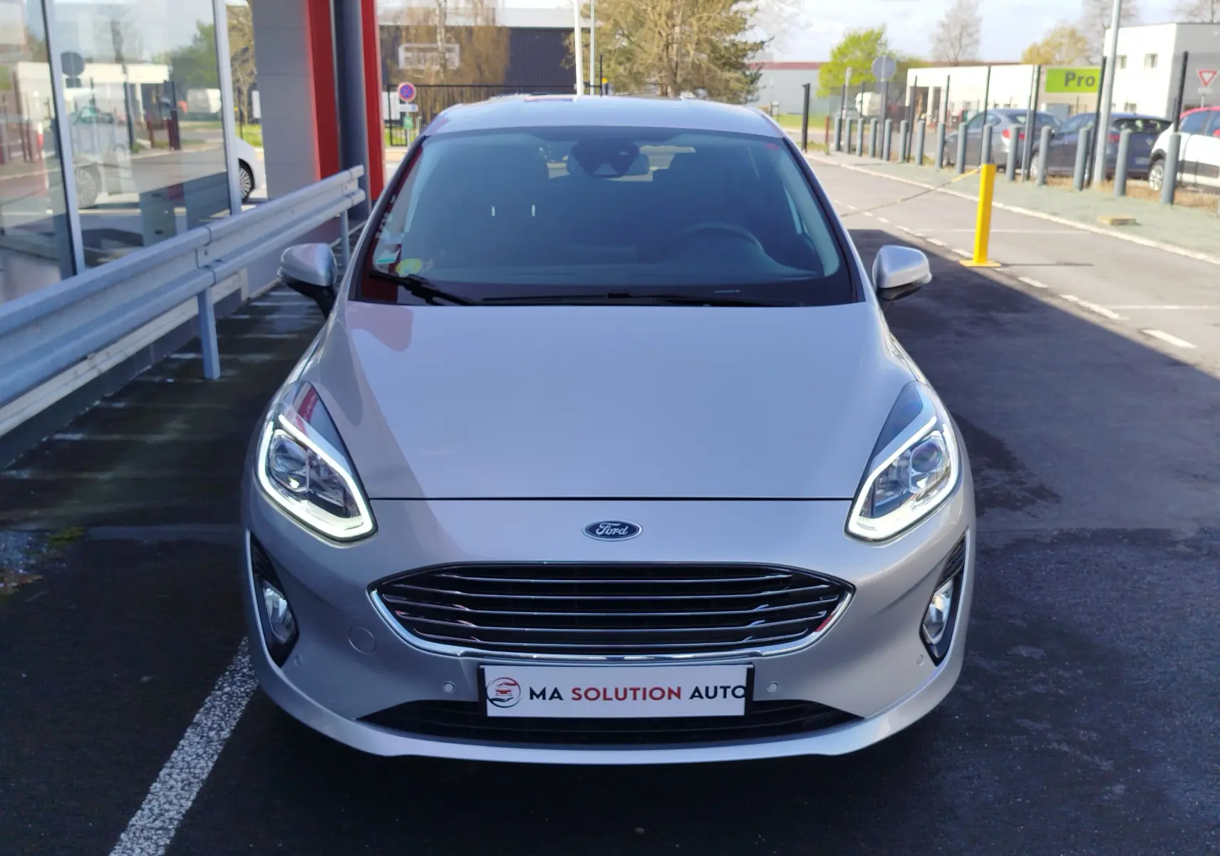 Vue avant d'une Ford Fiesta gris lunaire 2019 avec phares LED allumés, sur un parking extérieur.
