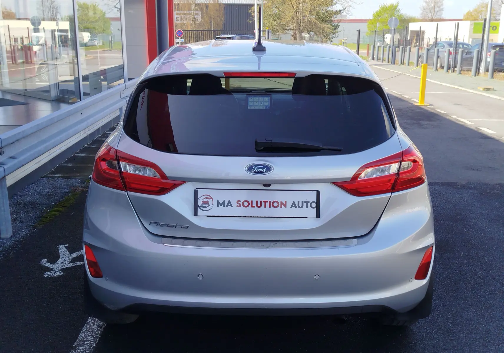 Vue arrière d'une Ford Fiesta gris lunaire 5 portes, avec feux arrière rouges et vitres teintées, stationnée en extérieur.