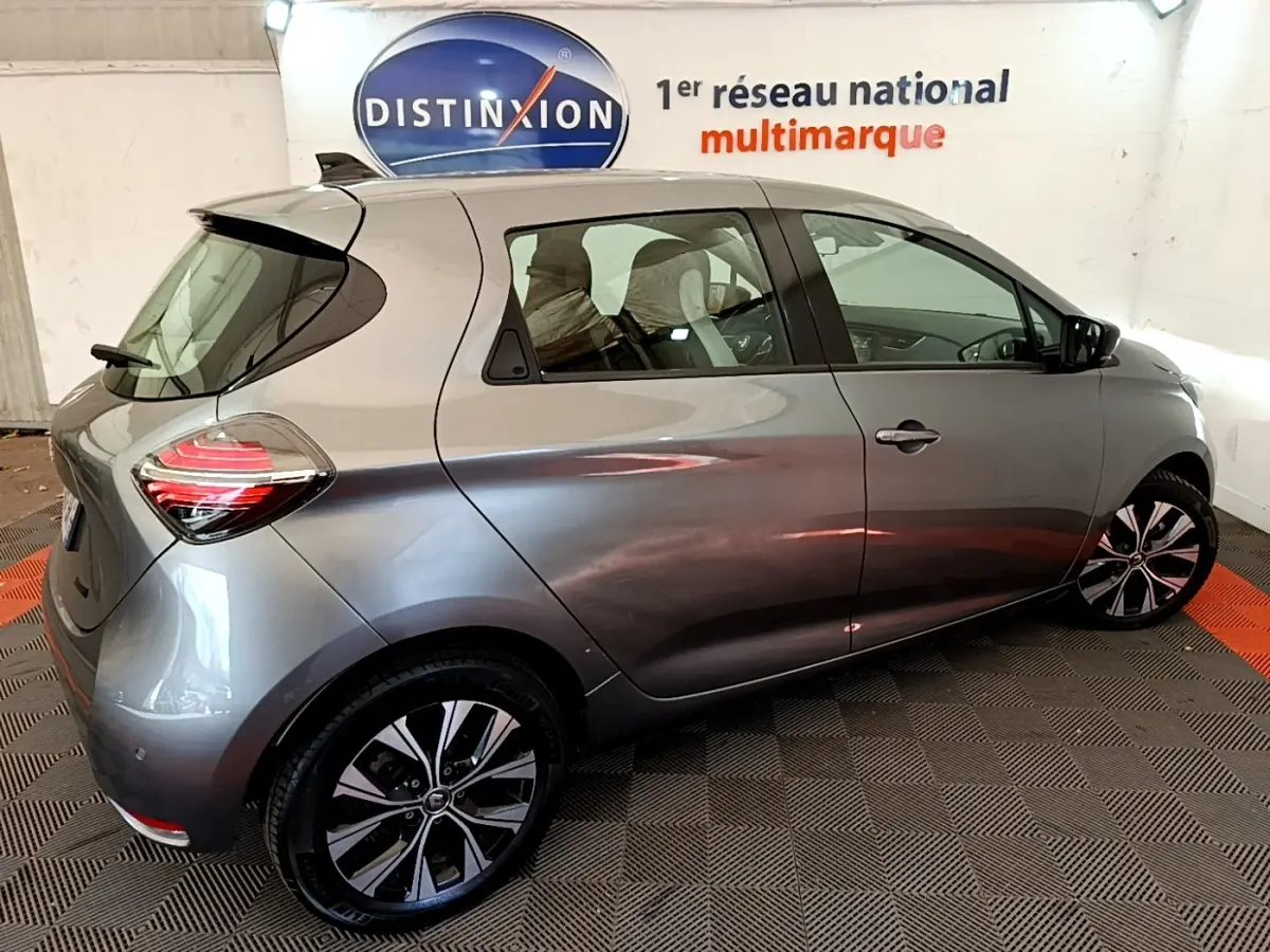 Vue 3/4 arrière droite d'une Renault ZOE gris clair électrique, avec jantes noires et logo Renault visible sur la roue.