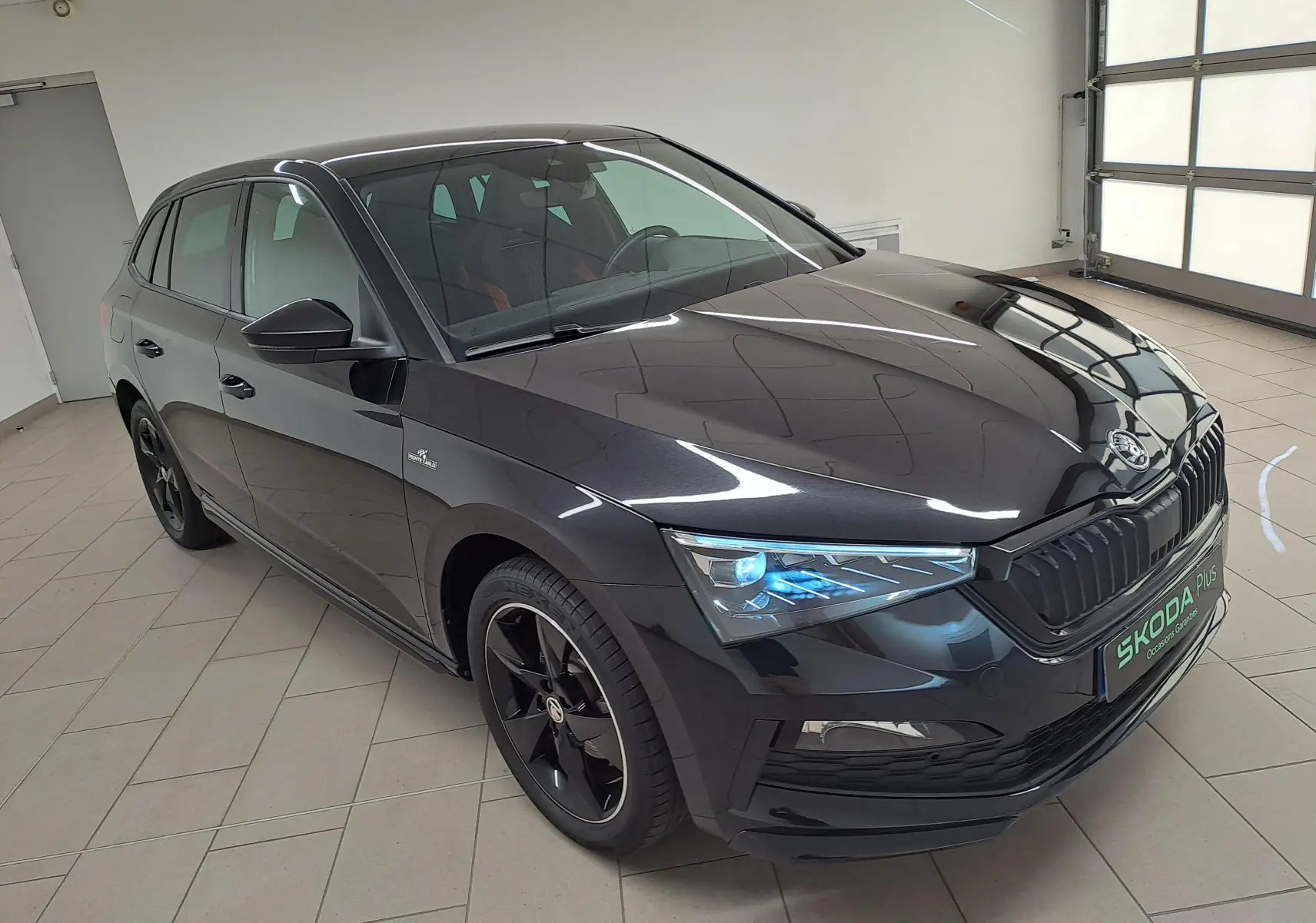 Vue 3/4 avant droite d'une Skoda Scala 1.0 TSI Monte-Carlo noire nacrée avec jantes noires et phares LED allumés.