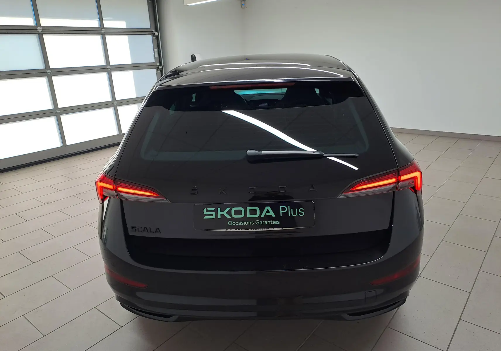 Vue arrière d'une Skoda Scala 2022 en Noir Magic nacré avec feux arrière allumés dans un garage lumineux.