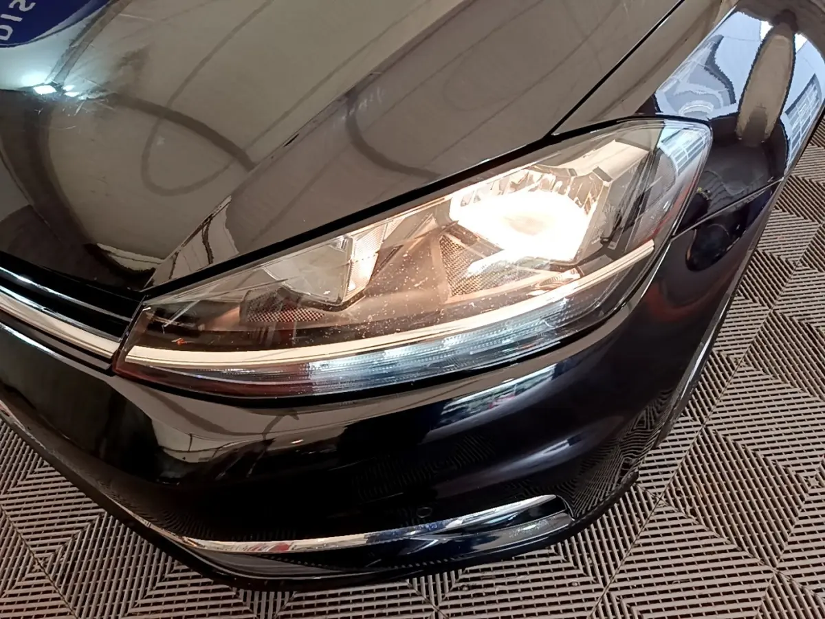Gros plan sur le phare avant droit allumé d’une Volkswagen Golf noire, avec calandre chromée visible.