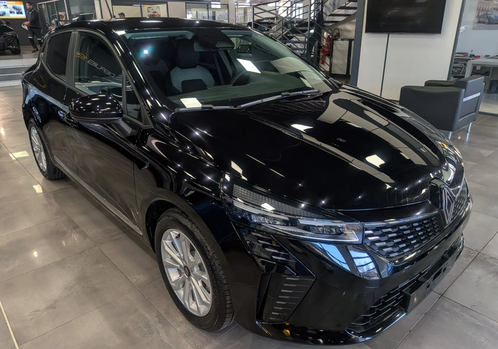 Vue 3/4 avant droite d'une Renault Clio V noire 2025 en intérieur, avec calandre et phares LED distinctifs.