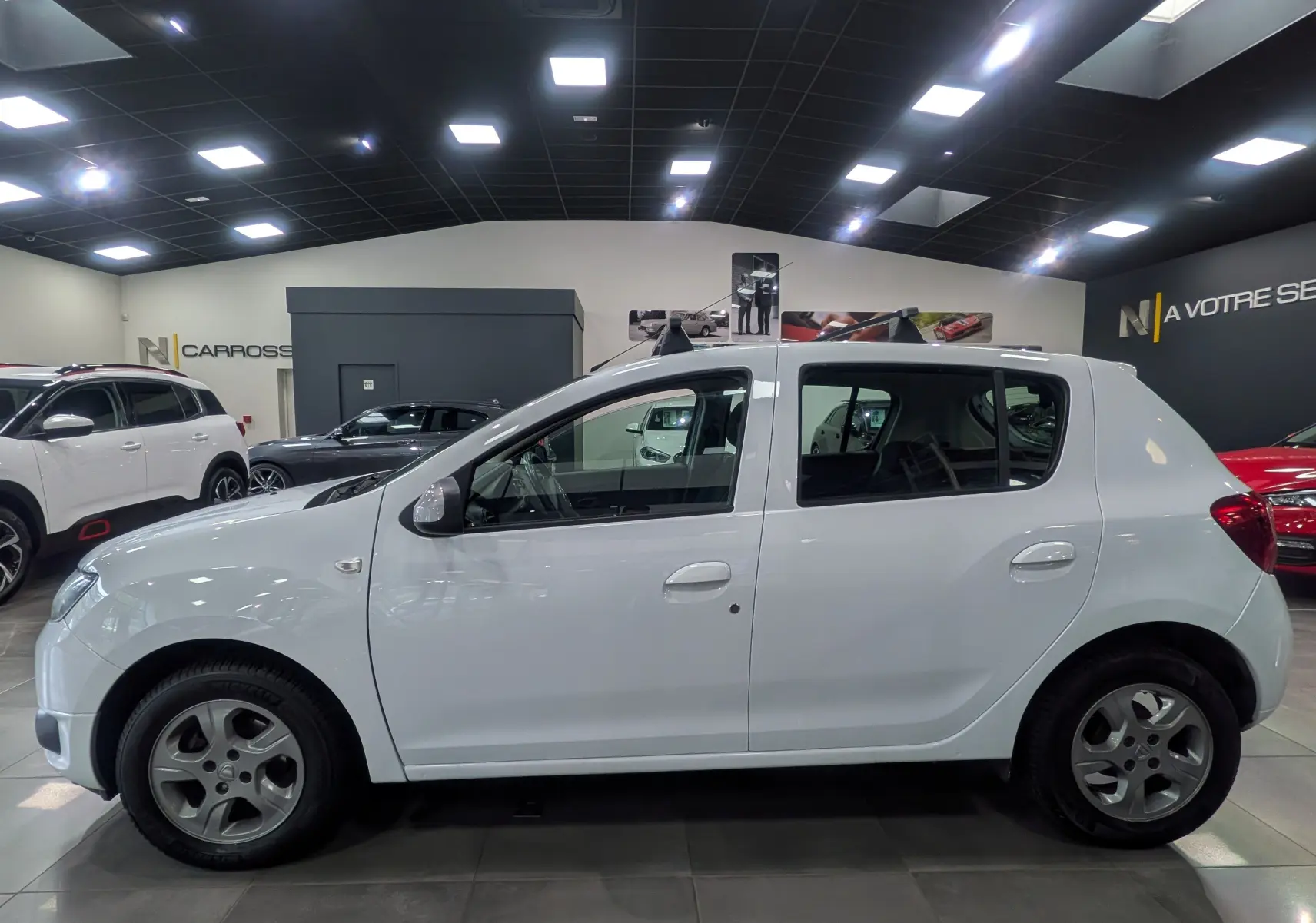 Profil côté droit d'une Dacia Sandero blanc glacier de 2015 en intérieur, avec ses lignes simples et ses vitres teintées.