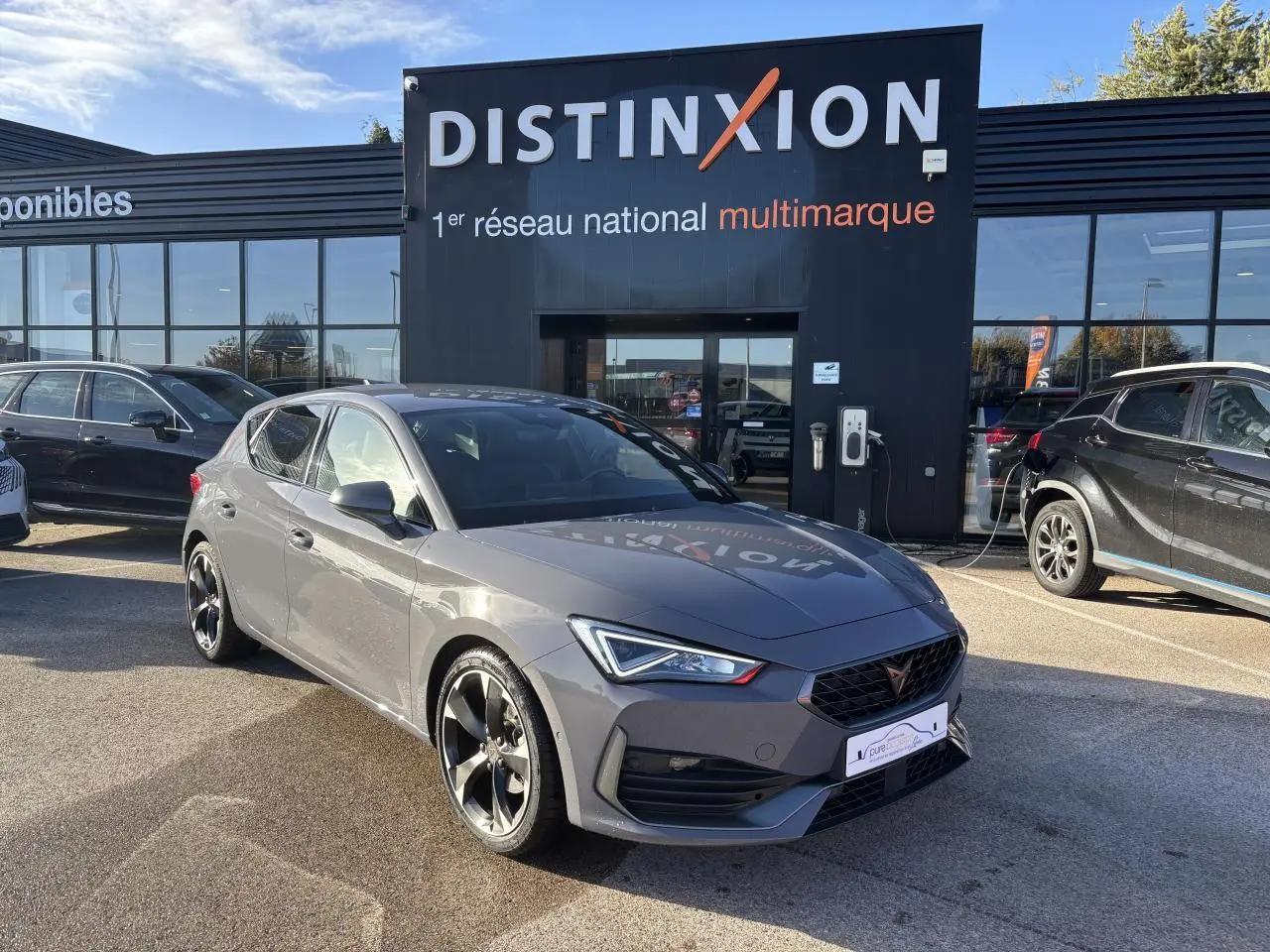 Vue 3/4 avant d'une CUPRA Leon gris Graphene avec jantes alliage noires devant un showroom Distinxion.