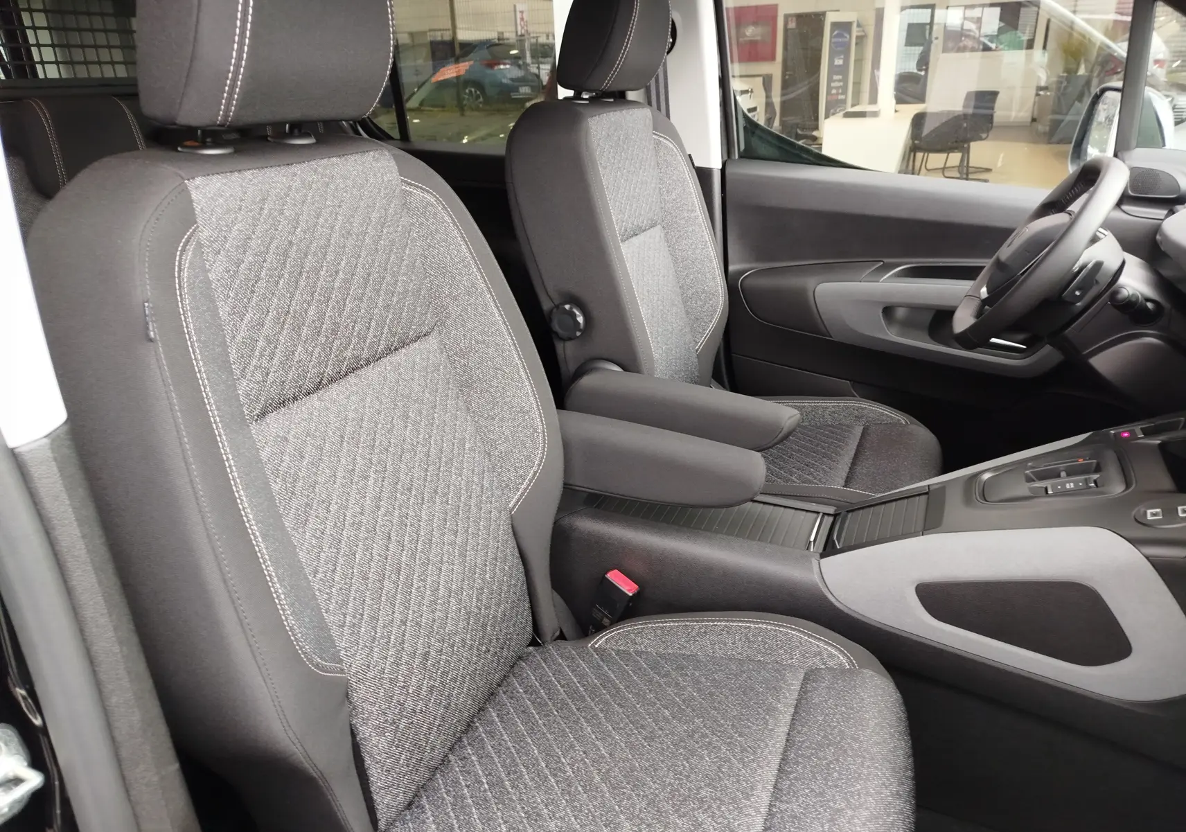 Vue intérieure côté conducteur du Peugeot Rifter 2026, sièges tissu gris avec accoudoir central et volant visible.
