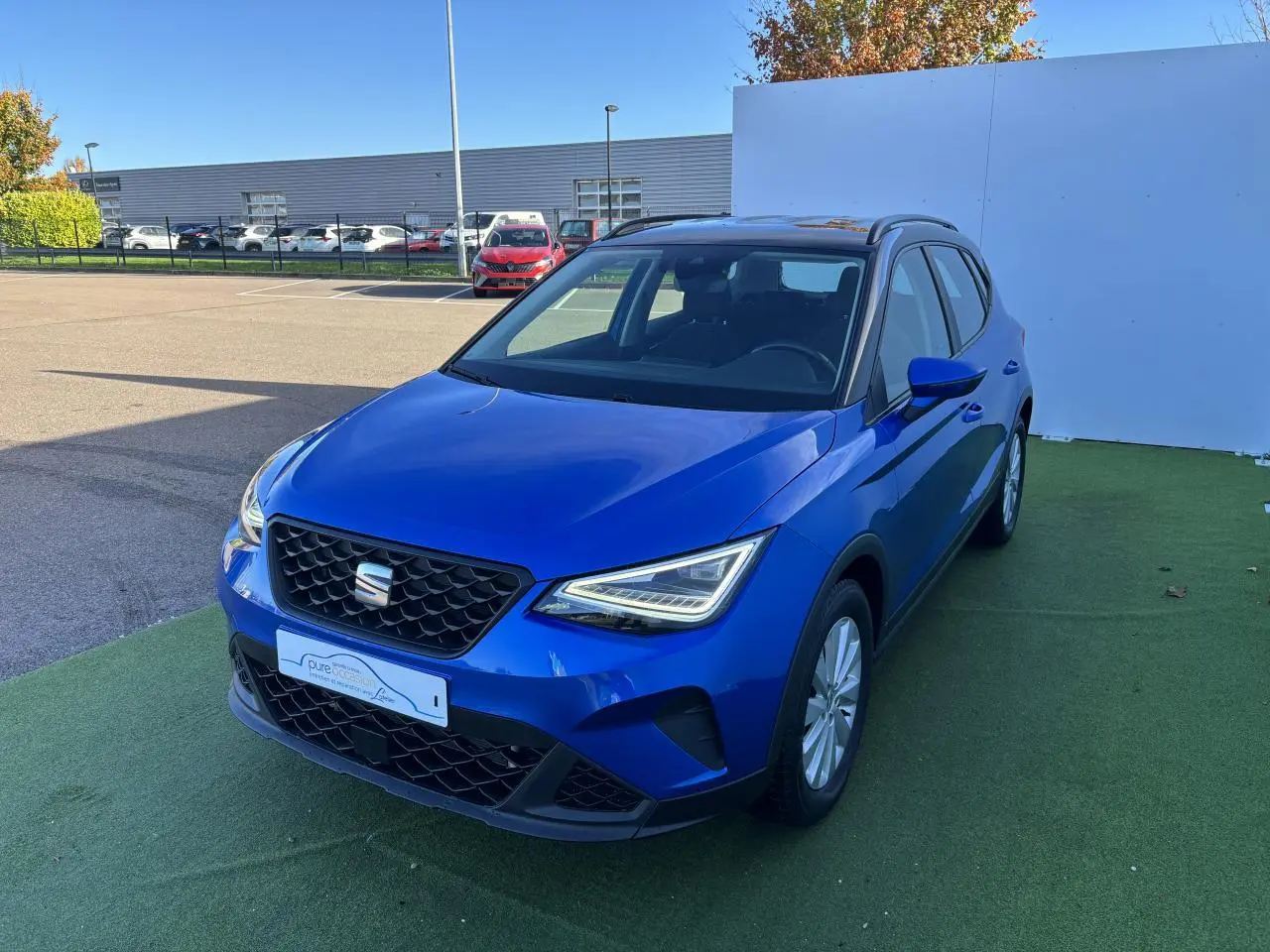 Vue 3/4 avant d'un SEAT Arona 2024 bleu saphir avec toit noir et jantes alliage, stationné sur sol vert.