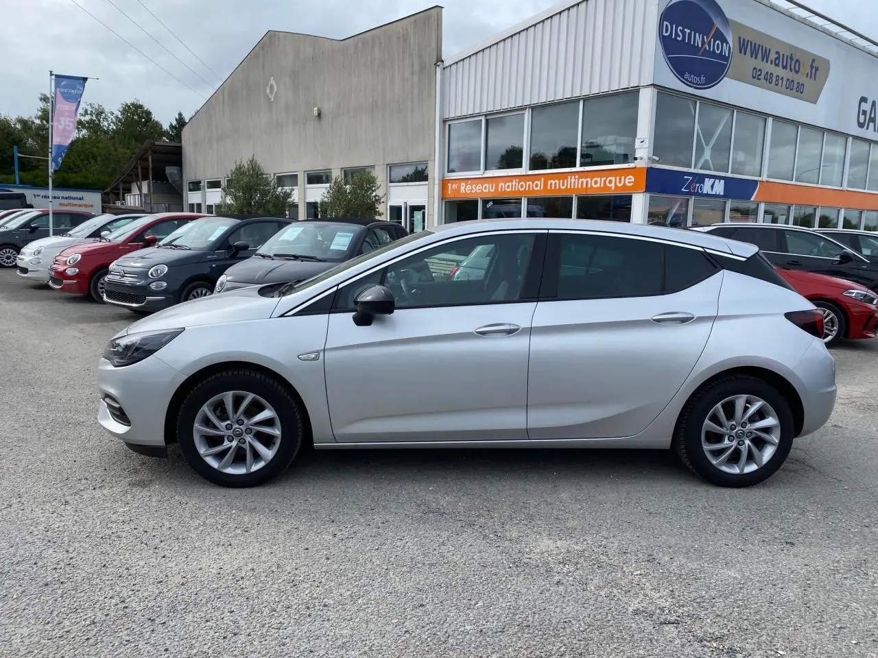 Profil côté gauche d'une Opel Astra Sovereign Silver 2021 garée devant un garage multimarque avec jantes alliage 16 pouces.