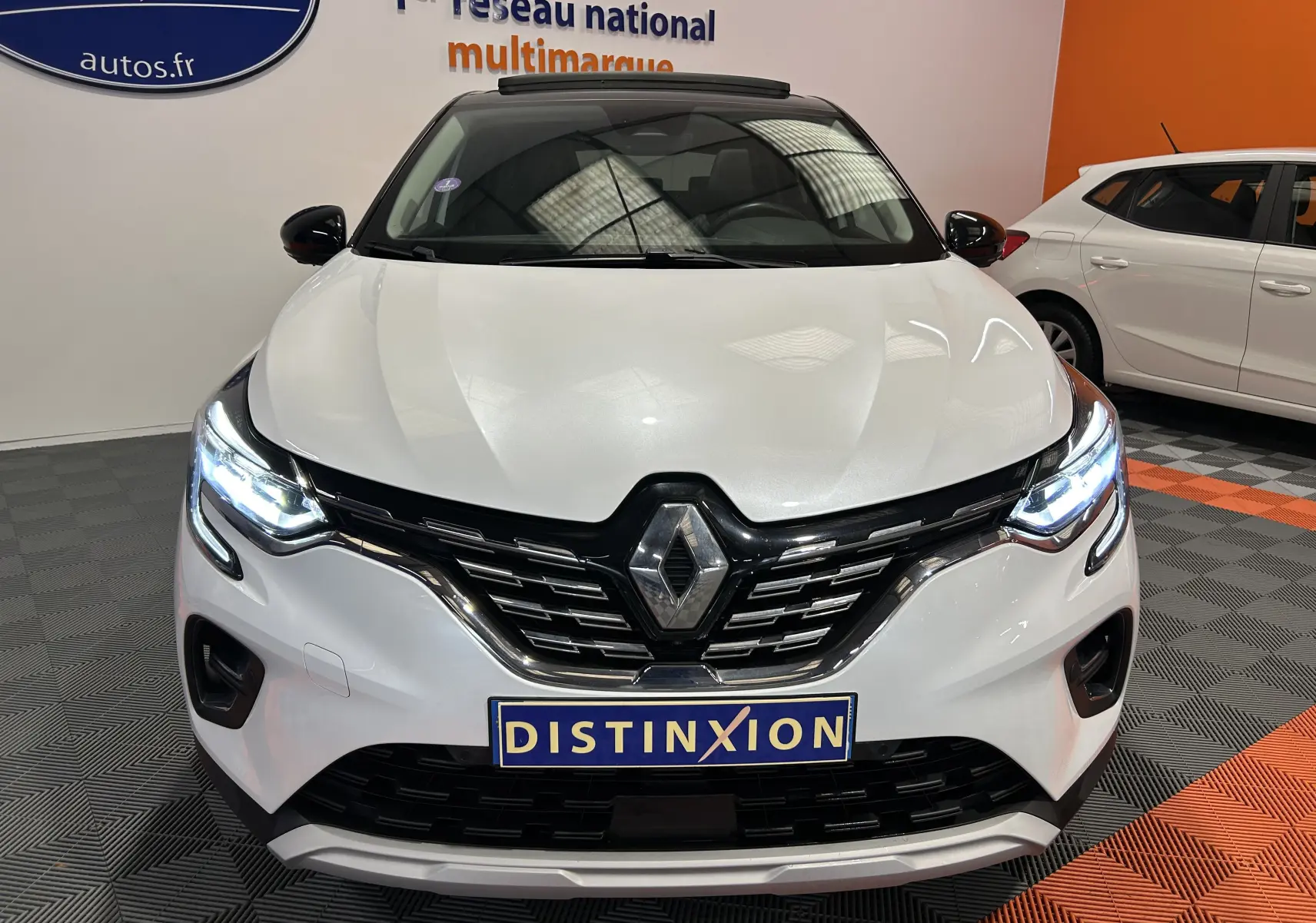 Vue frontale d'un Renault Captur blanc nacré 2022 avec phares allumés et toit ouvrant visible en intérieur showroom.