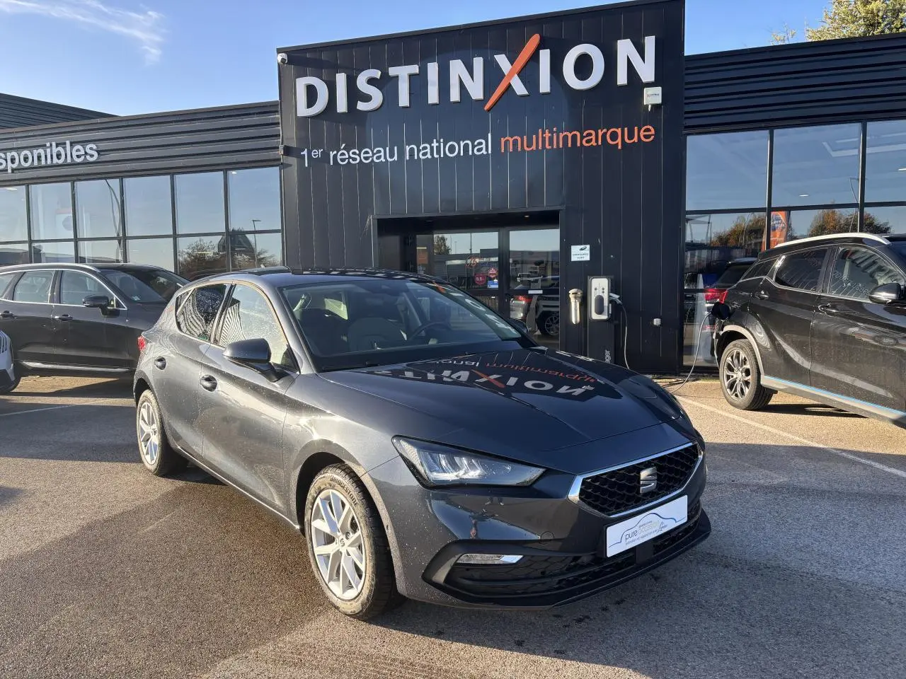 SEAT Leon IV gris magnetic vue 3/4 avant droit, jantes alliage 16 pouces, stationnée devant un showroom Distinxion.