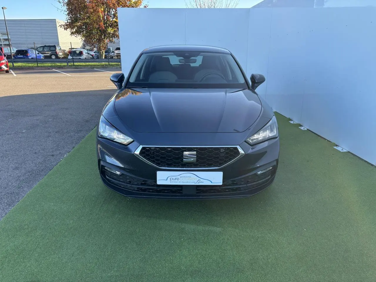 Vue frontale d'une SEAT Leon IV gris magnetic garée sur un sol vert avec calandre hexagonale et phares LED allumés.