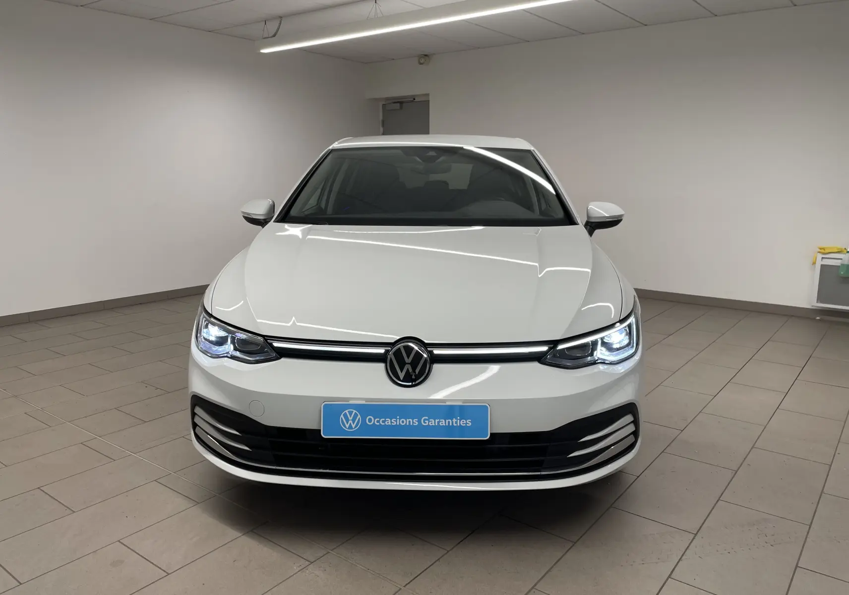 Volkswagen Golf 1.5 eTSI blanc pur vue de face en intérieur avec phares allumés et logo VW central.