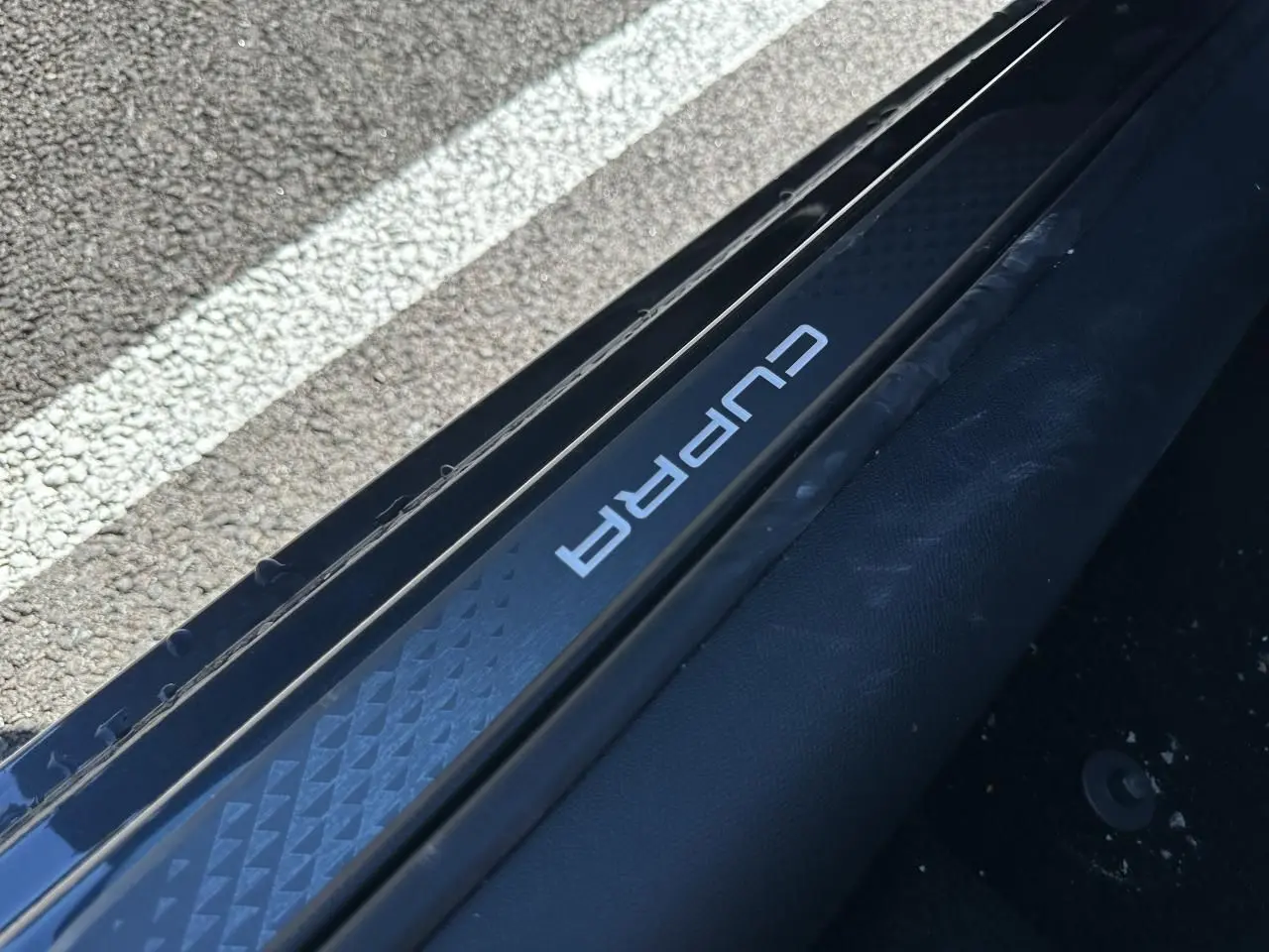 Gros plan sur le seuil de porte côté gauche avec logo CUPRA sur fond noir texturé, intérieur sombre visible.