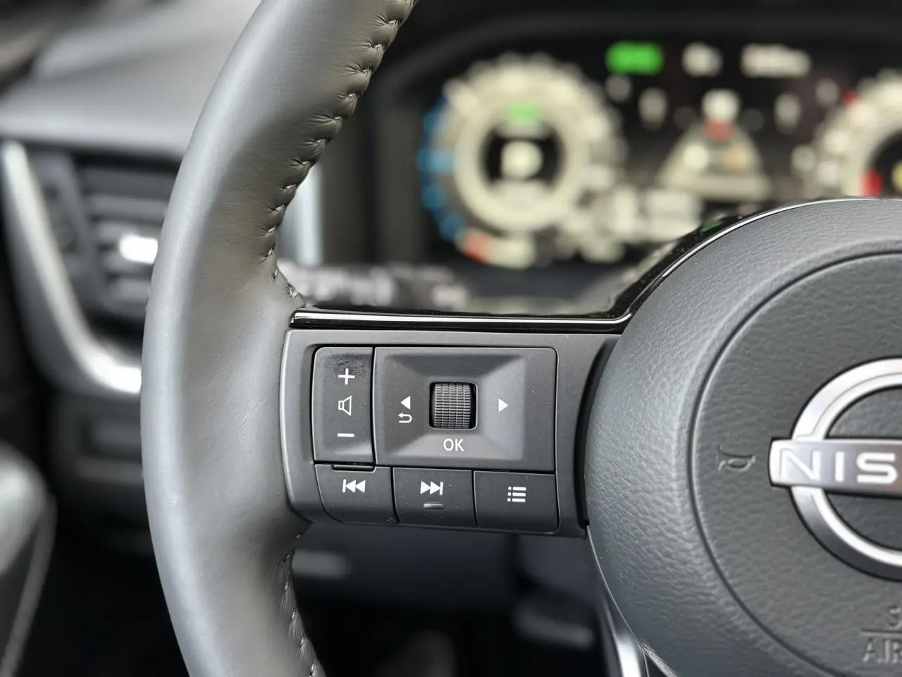 Gros plan sur les commandes gauche du volant cuir gris du Nissan Qashqai III e-Power avec tableau de bord numérique flou en arrière-plan.