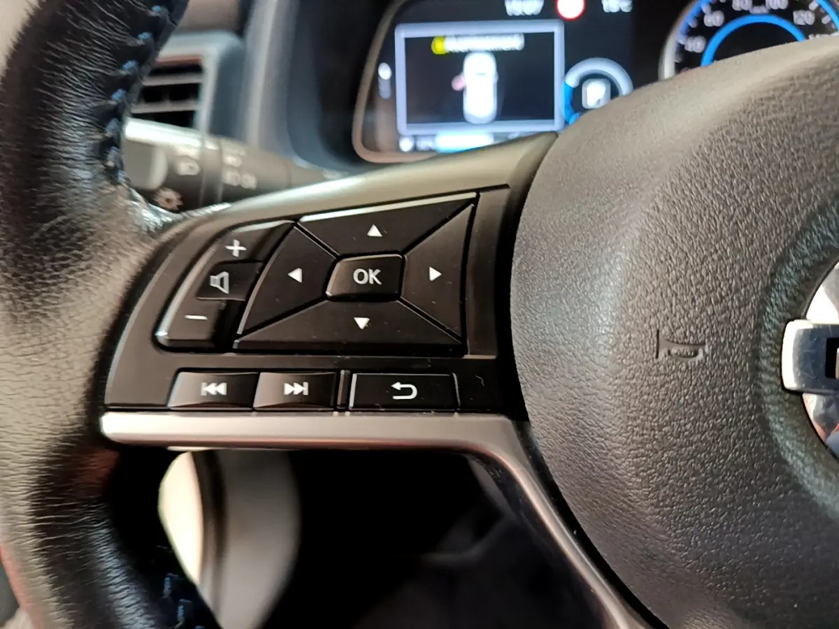 Gros plan sur les commandes gauche du volant cuir noir du Nissan Leaf blanc, avec affichage numérique flou en arrière-plan.