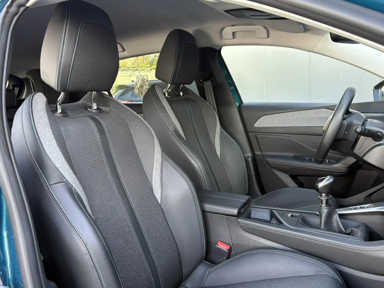 Vue intérieure côté conducteur de la Peugeot 308 2024, sièges tri matières noirs avec surpiqûres bleues et levier de vitesses manuel.