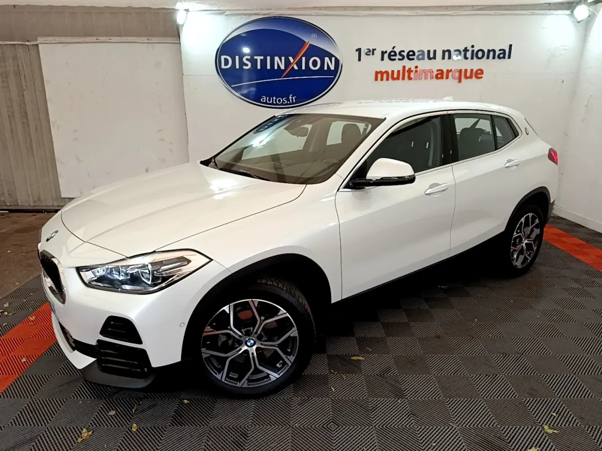 BMW X2 blanc en 3/4 avant droit dans un showroom, mettant en valeur ses phares LED et jantes design.