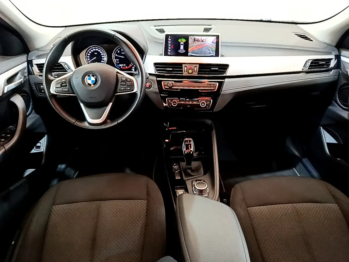 Vue intérieure frontale du tableau de bord et volant cuir noir de la BMW X2 sDrive18i 2023 avec écran central affichant l'aide au stationnement.