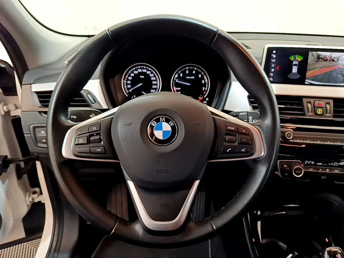 Vue intérieure centrée sur le volant cuir de la BMW X2 blanc, avec tableau de bord et écran d'aide au stationnement visibles.