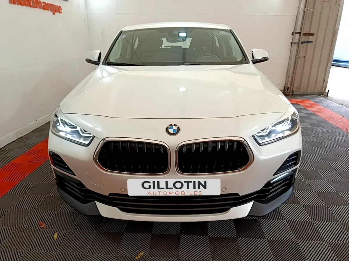 Vue de face d'un BMW X2 blanc avec calandre double haricot et phares LED allumés dans un garage.