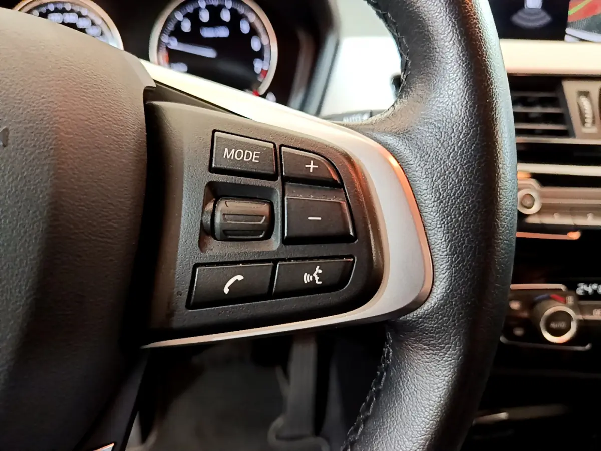 Gros plan sur les commandes du volant cuir noir de la BMW X2 2023, avec tableau de bord et écran en arrière-plan.