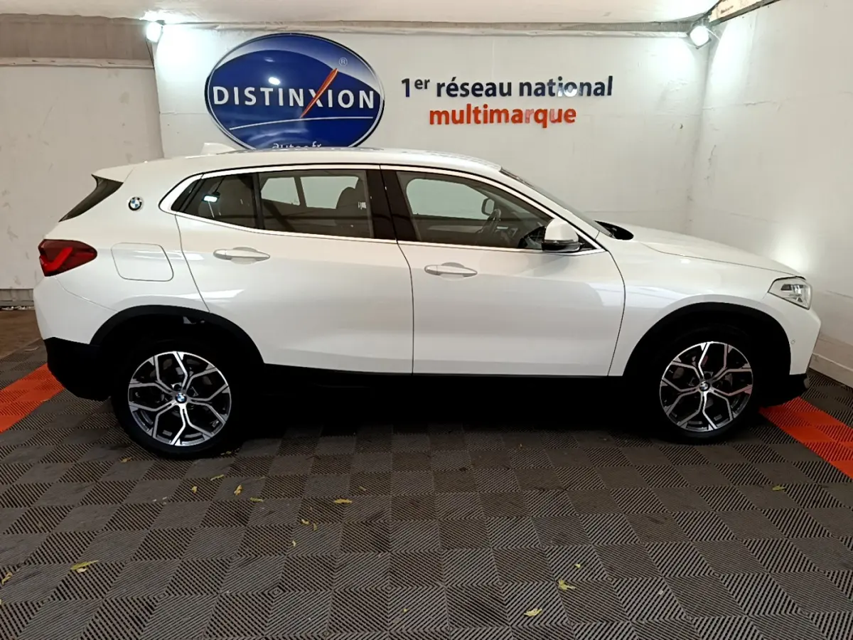 BMW X2 blanc vue de profil côté gauche dans un showroom avec jantes alliage et logo sur l'aile arrière.