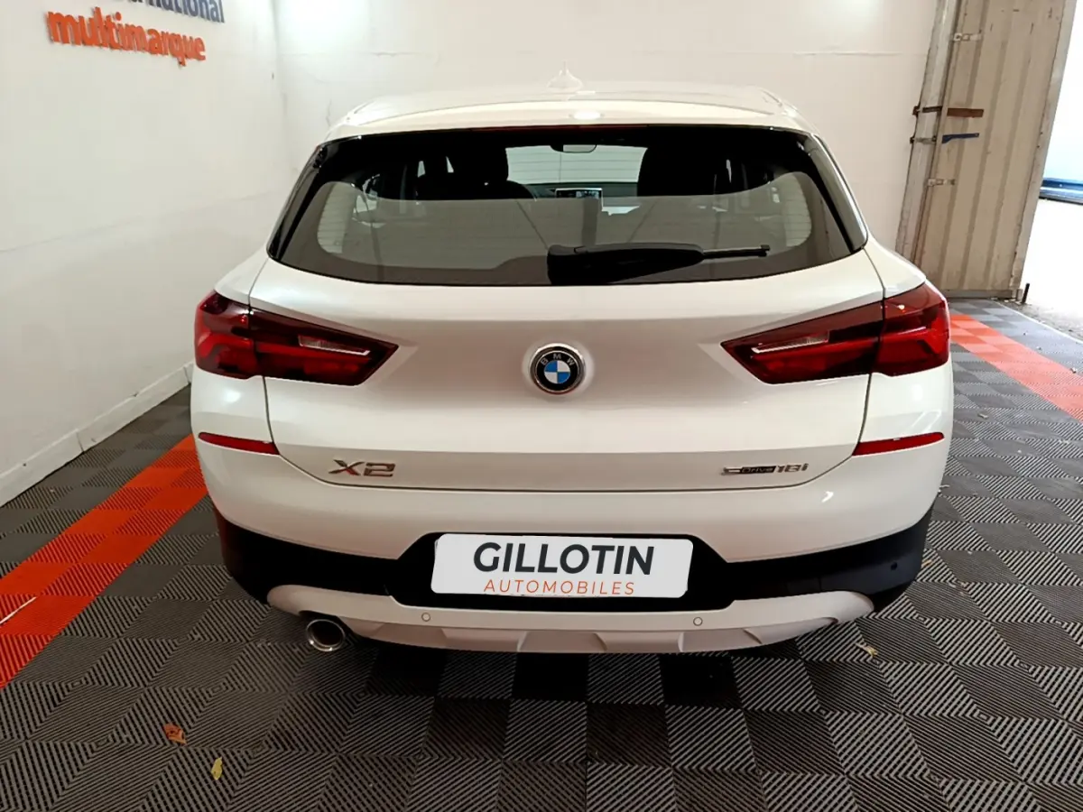 Vue arrière d'un BMW X2 blanc sDrive18i Business DesignDKG7 avec feux LED et sortie d'échappement visible.