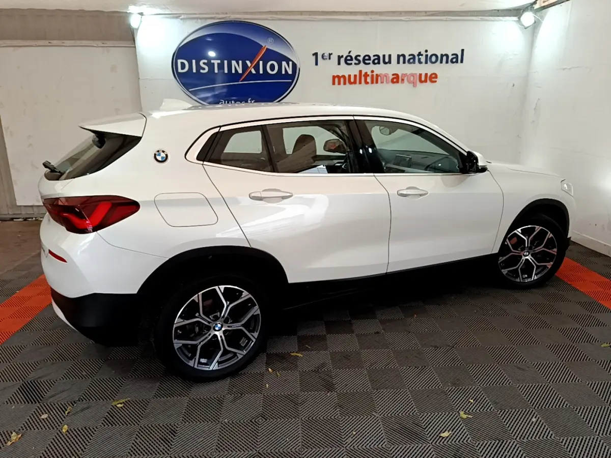 BMW X2 blanc vu de profil côté gauche dans un showroom avec jantes alliage et logo distinctif sur l'aile arrière.