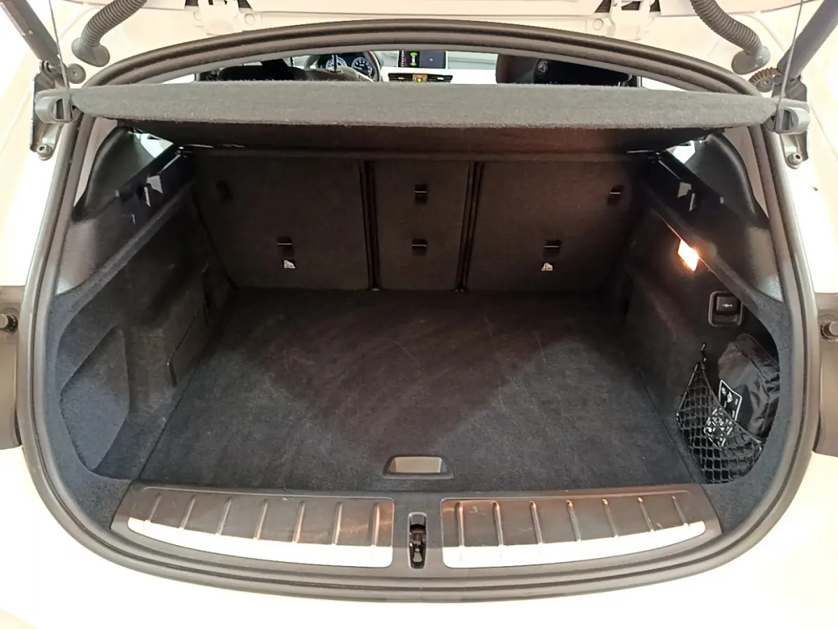 Coffre ouvert vu de l'arrière d'un BMW X2 blanc, avec tapis noir et éclairage intérieur à droite.