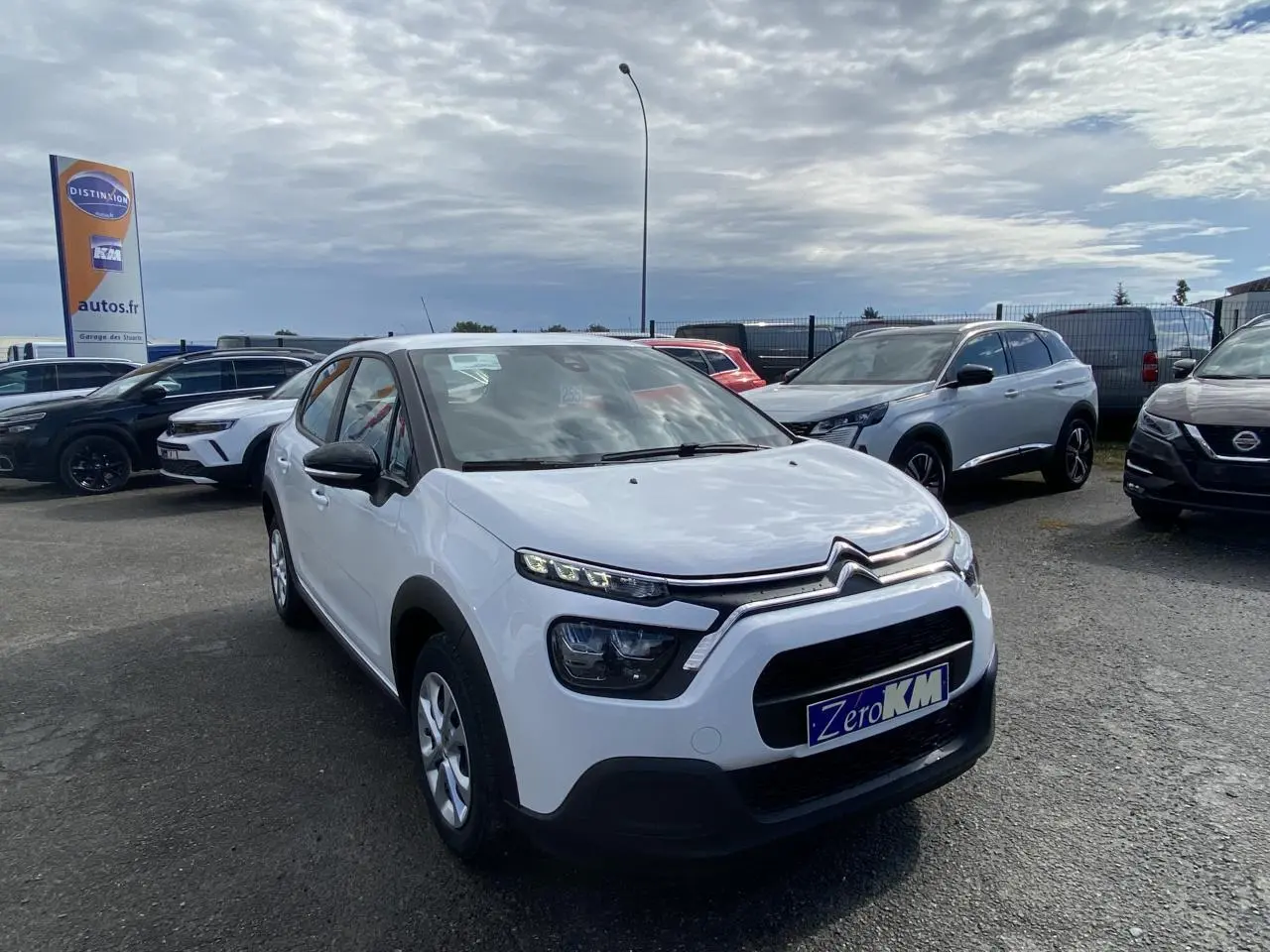 Citroën C3 blanc vue 3/4 avant droit, avec calandre noire et enjoliveurs 15 pouces sur parking extérieur.
