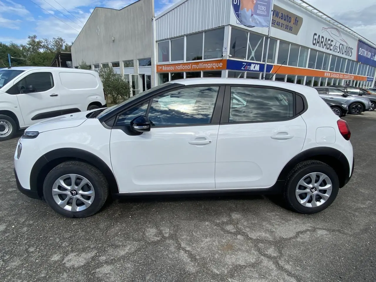 Profil côté gauche d'une Citroën C3 blanche 2022 avec enjoliveurs 15 pouces et rétroviseurs noirs devant un garage.