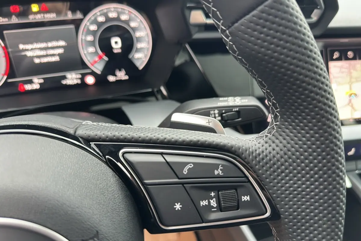 Gros plan sur le volant cuir perforé de l'Audi A3 2025 avec commandes audio et palettes au volant visibles.