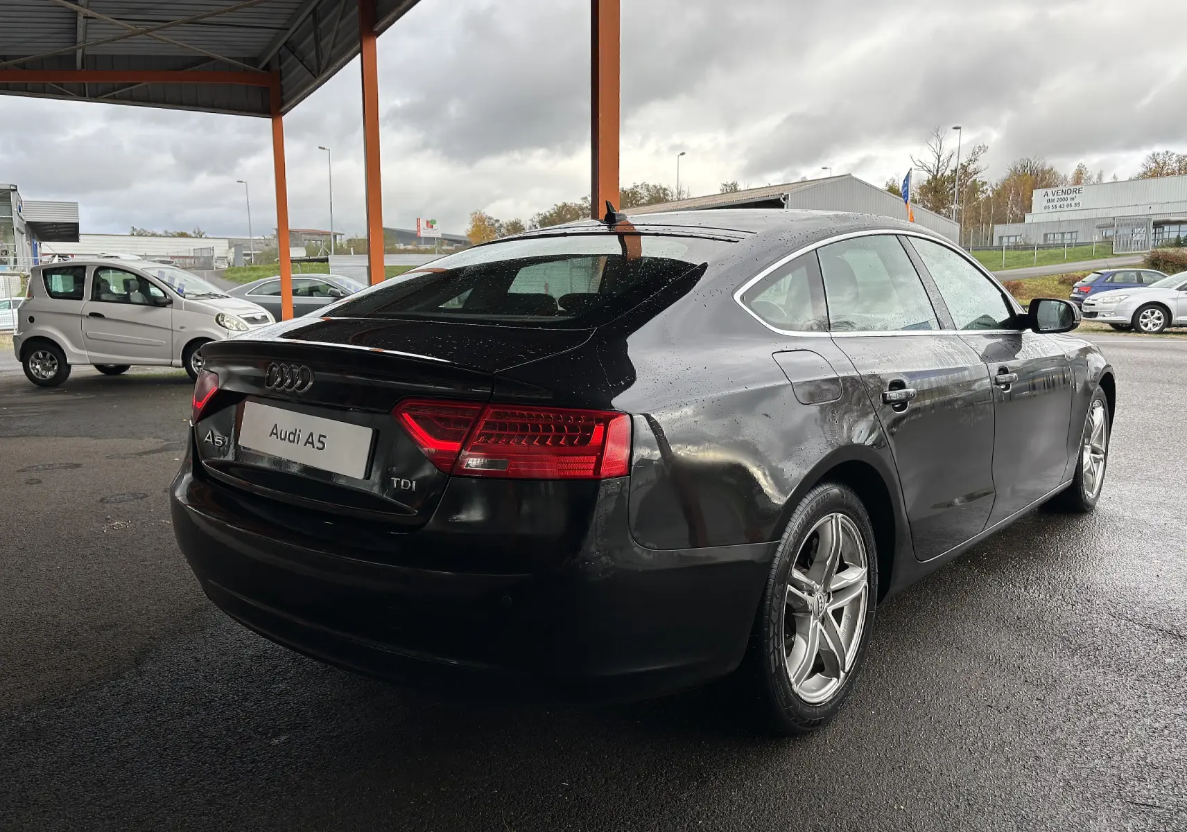 Audi A5 Sportback noir vue 3/4 arrière droit sous abri avec feux arrière allumés par temps couvert.