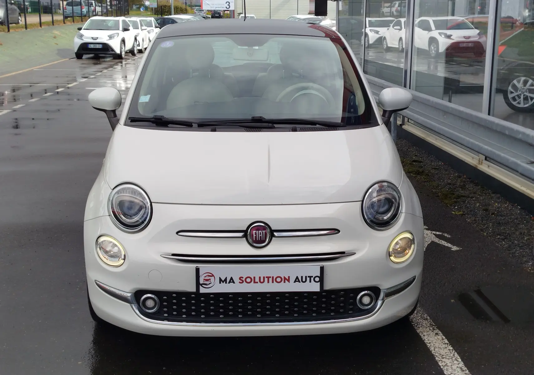 Vue frontale d'une Fiat 500 blanche Bossa Nova, avec ses phares ronds caractéristiques allumés, stationnée devant un bâtiment vitré.