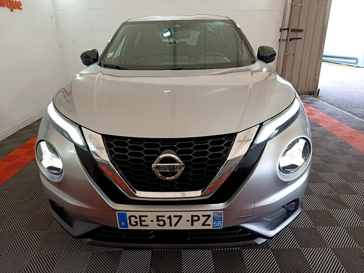 Vue avant d'un Nissan Juke gris clair 2022 avec phares LED allumés et calandre chromée en intérieur garage.