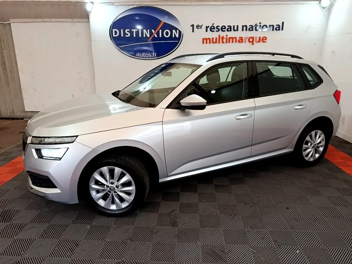 Vue de profil côté gauche d'un Skoda Kamiq gris clair 2022 en showroom avec jantes alliage et barres de toit noires.