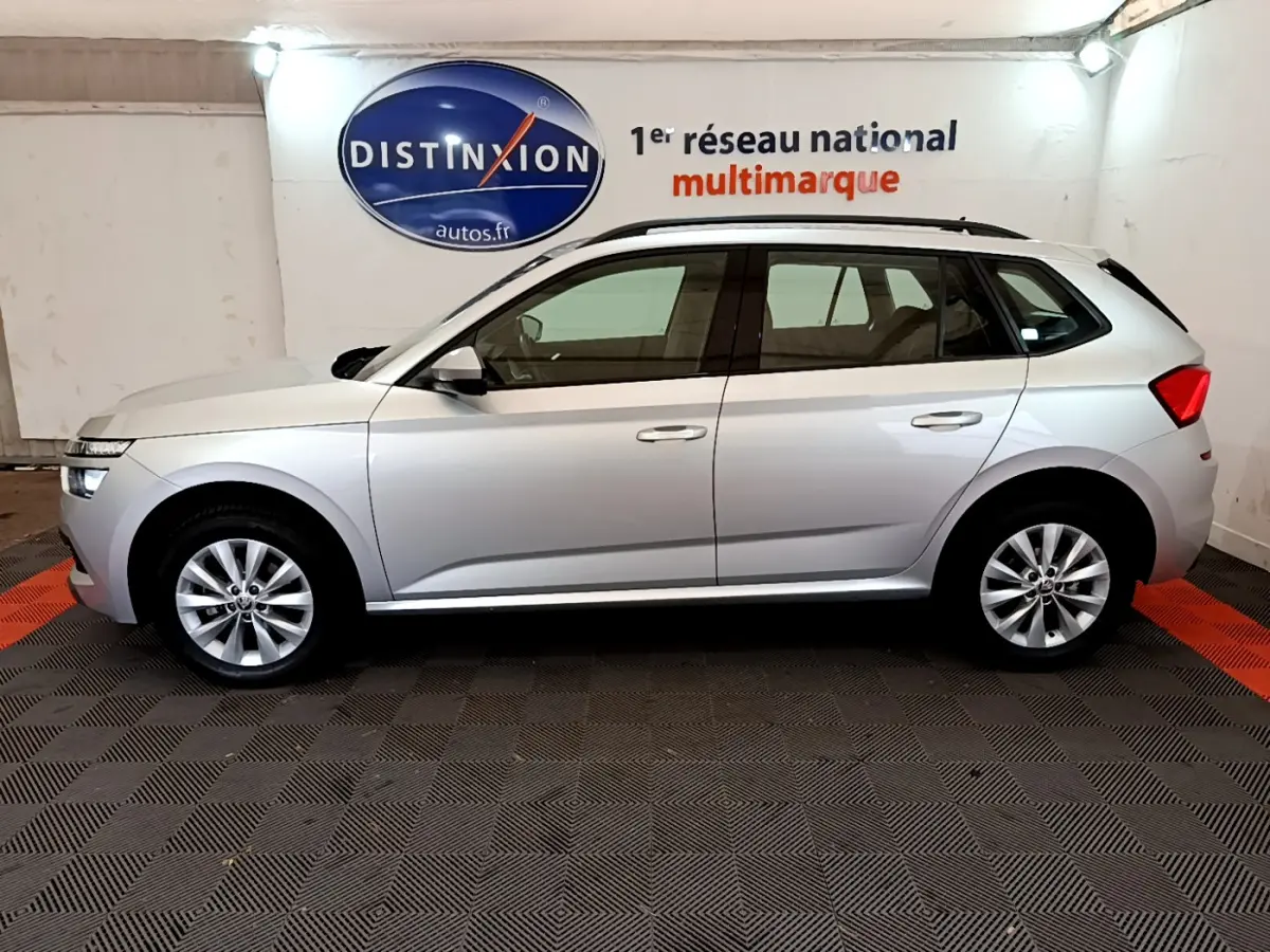Vue de profil côté gauche d'un Skoda Kamiq gris clair 2022 dans un showroom avec logo Distinxion en arrière-plan.