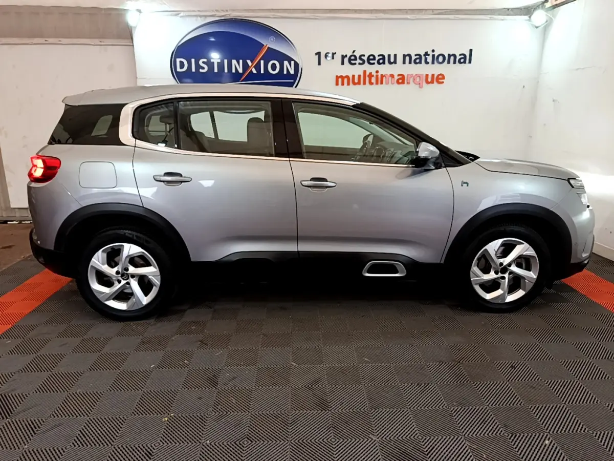 Profil côté droit du Citroën C5 Aircross Hybrid gris clair avec jantes alliage et feux arrière allumés en intérieur showroom.