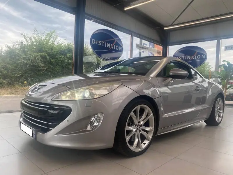 Peugeot RCZ gris en 3/4 avant droit, avec jantes alliage et phares allumés dans un showroom lumineux.