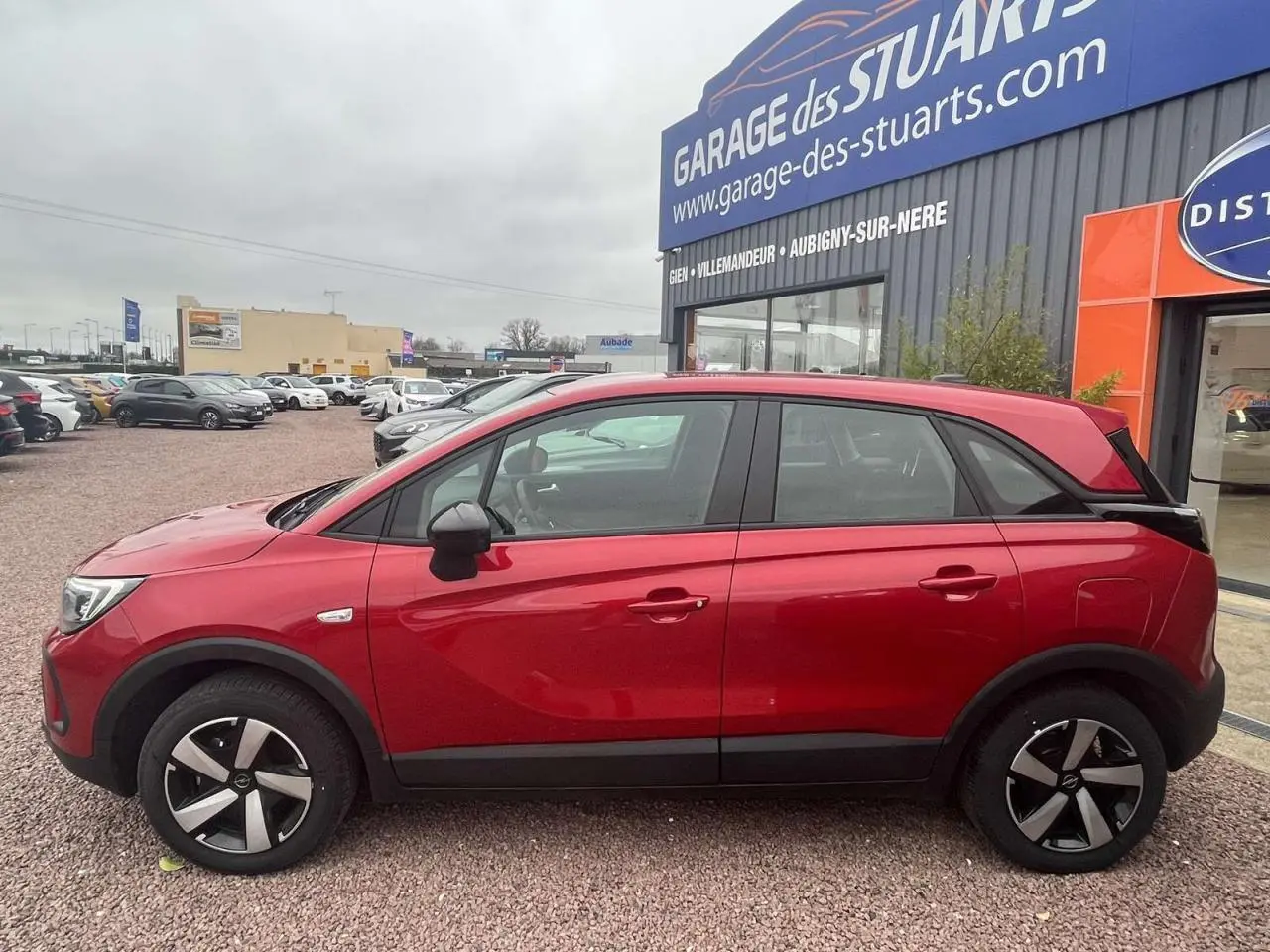Profil côté gauche d'un Opel Crossland rouge métallisé 2022 garé devant un garage avec jantes noires et argentées.